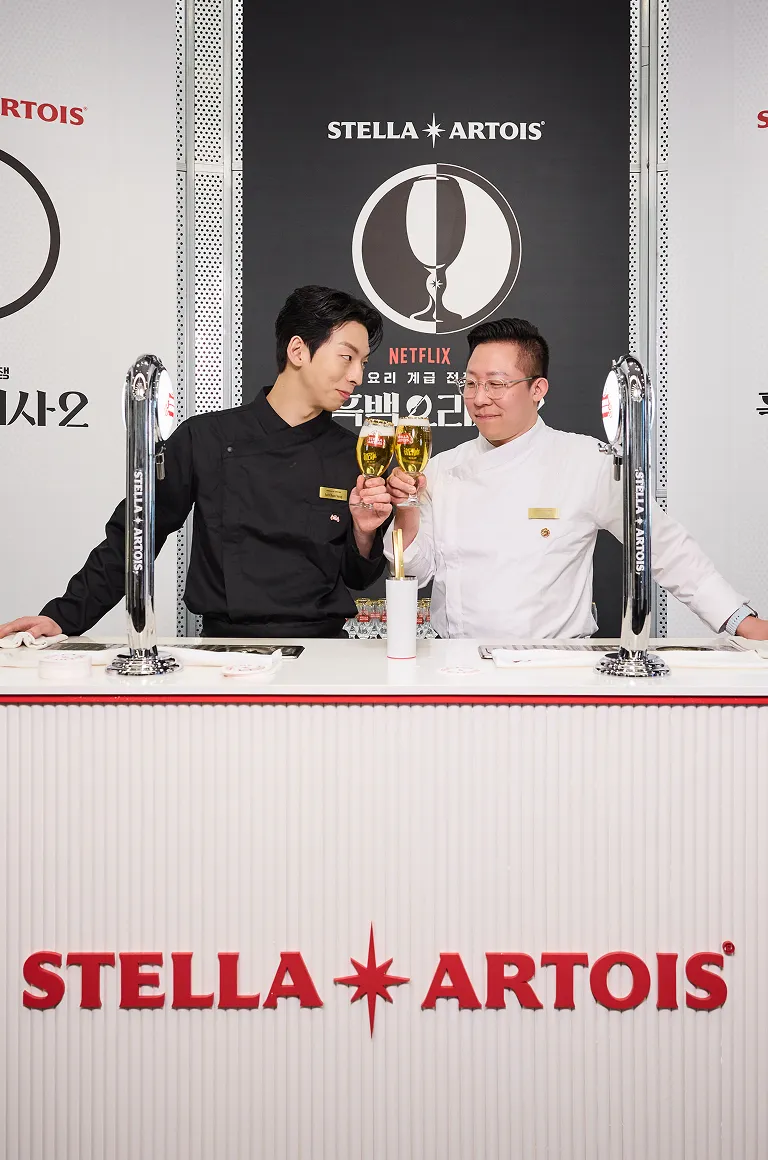 흰색 셰프복을 입은 이준 셰프와 검은색 셰프복을 입은 이찬양 셰프가 스텔라 아르투아 생맥주 잔을 들고 건배하고 있습니다