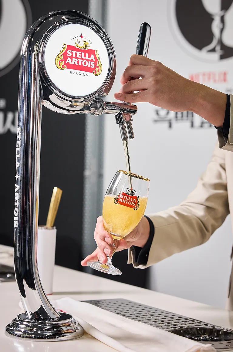 드래프트 마스터가 스텔라 아르투아 탭에서 황금빛 생맥주를 챌리스 잔에 따르고 있으며, 잔 위로 풍성한 거품이 올라오고 있습니다