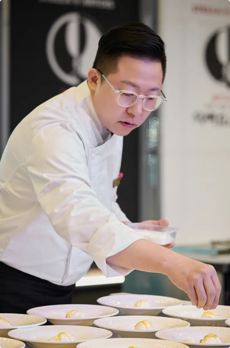 흰색 셰프복과 안경을 쓴 이준 셰프가 여러 개의 흰 접시 위에 디저트를 정성스럽게 플레이팅하고 있습니다