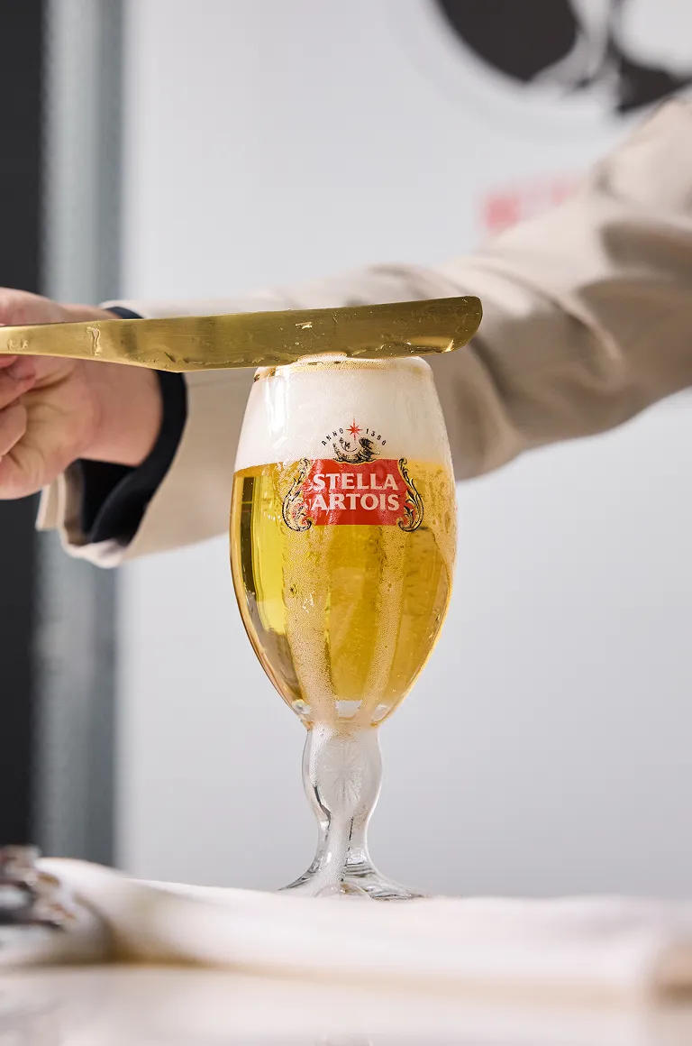 드래프트 마스터가 금색 트레이를 기울여 스텔라 아르투아 챌리스 잔의 거품을 깔끔하게 정리하고 있습니다