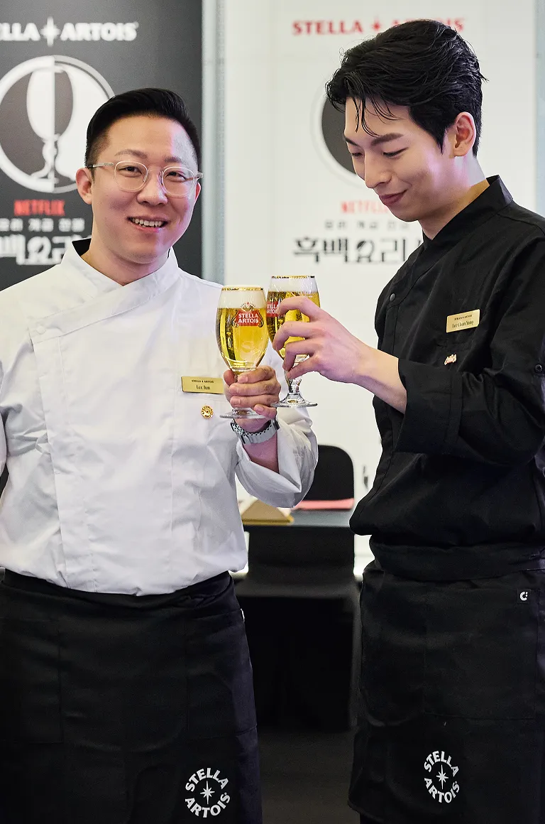 흰색 셰프복의 이준 셰프와 검은색 셰프복의 이찬양 셰프가 환하게 웃으며 스텔라 아르투아 생맥주 잔을 부딪히며 건배하고 있습니다