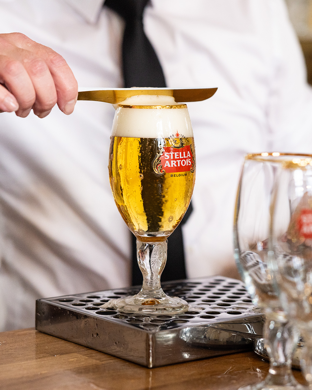 흰 셔츠와 검정 넥타이를 착용한 바텐더가 Stella Artois 로고가 새겨진 전용 글라스에 맥주 거품을 금속 스패출라로 정리하는 장면.
