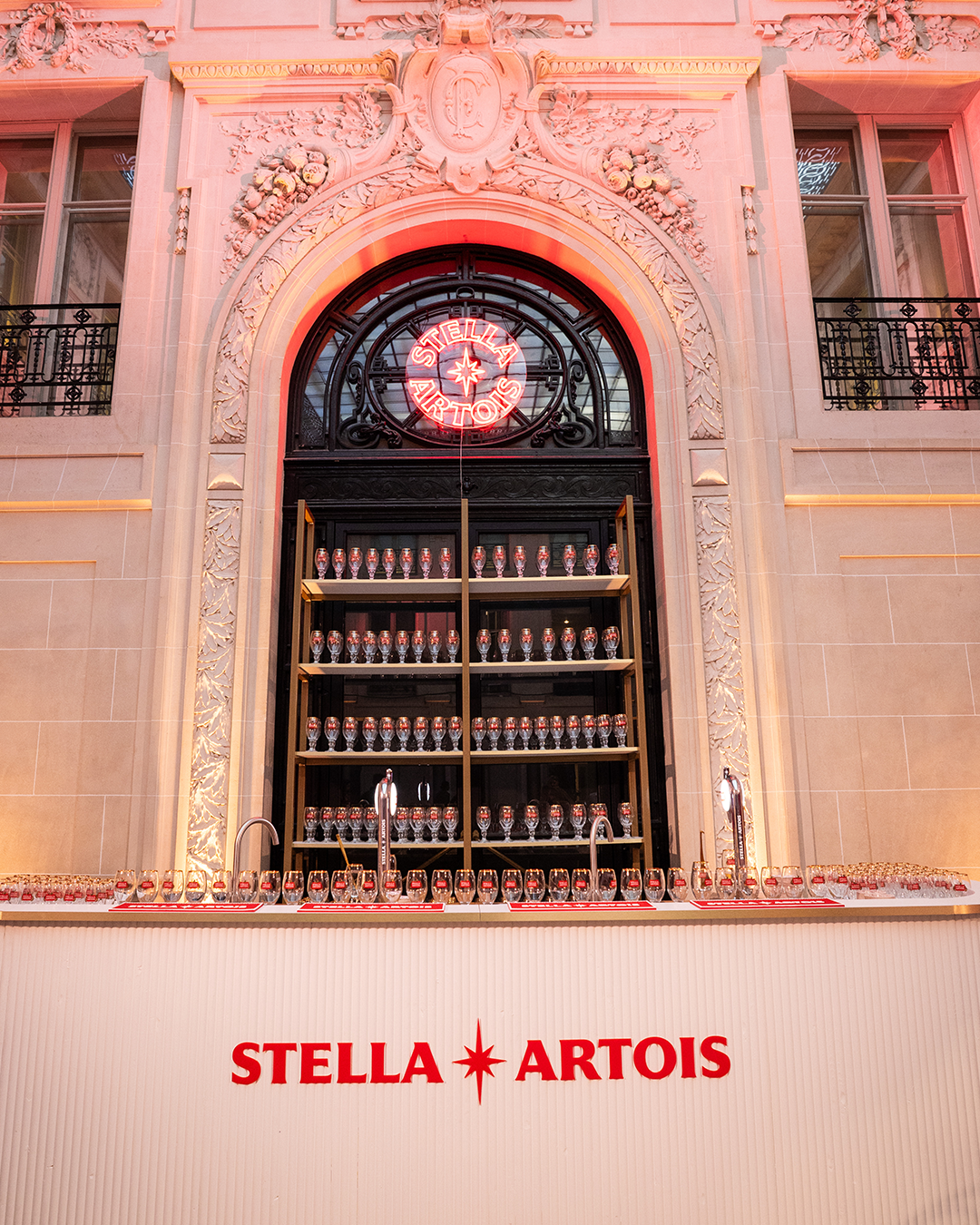 파리풍 클래식 건물 아치형 창문 앞에 설치된 Stella Artois 브랜드 바 부스. 선반에 브랜드 전용 잔이 가득 진열되어 있고, 창문 상단에는 네온사인 로고가 빛나고 있다.