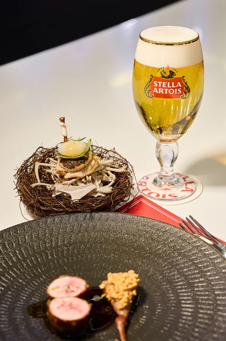 나뭇가지 장식 위에 올려진 메추리 요리와 검은 접시 위 메추리 다리 요리가 있고, 그 옆에 황금빛 스텔라 아르투아 생맥주가 담긴 챌리스 잔이 놓여 있습니다