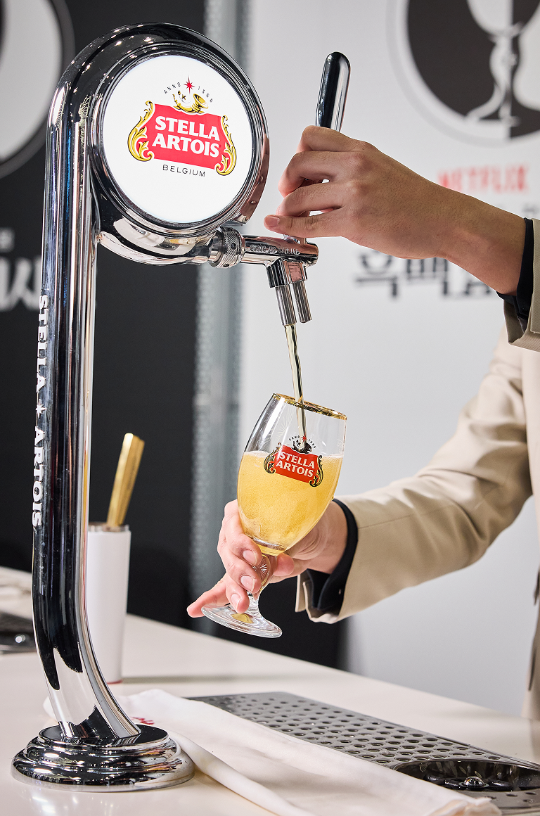 드래프트 마스터가 스텔라 아르투아 탭에서 황금빛 생맥주를 챌리스 잔에 따르고 있으며, 잔 위로 풍성한 거품이 올라오고 있습니다