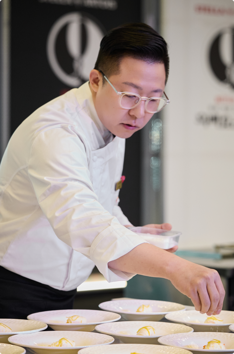 흰색 셰프복과 안경을 쓴 이준 셰프가 여러 개의 흰 접시 위에 디저트를 정성스럽게 플레이팅하고 있습니다