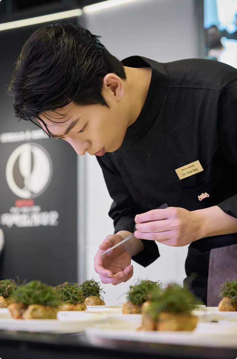 검은색 셰프복을 입은 이찬양 셰프가 고개를 숙이고 접시 위 요리에 허브를 올리며 집중해서 플레이팅하고 있습니다