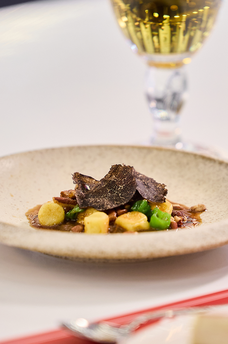 베이지색 접시 위에 소꼬리 조림이 담겨 있고, 그 위에 검은 트러플 슬라이스가 올려져 있으며 뒤로 와인잔이 보입니다