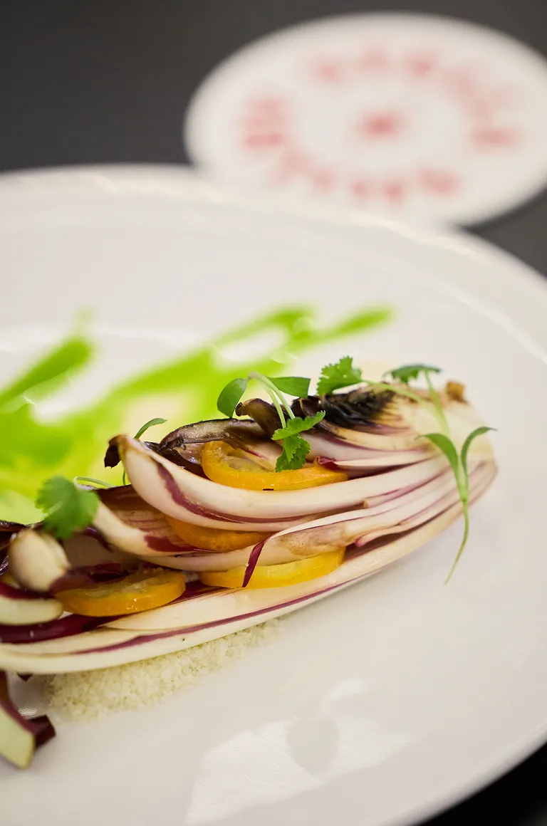 흰 접시 위에 보라색 타르디보와 녹색 소스가 곁들여진 치즈 요리가 예술적으로 플레이팅되어 있습니다