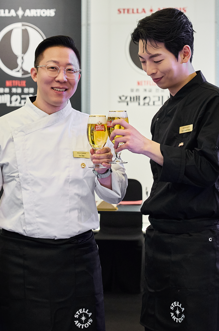 흰색 셰프복의 이준 셰프와 검은색 셰프복의 이찬양 셰프가 환하게 웃으며 스텔라 아르투아 생맥주 잔을 부딪히며 건배하고 있습니다