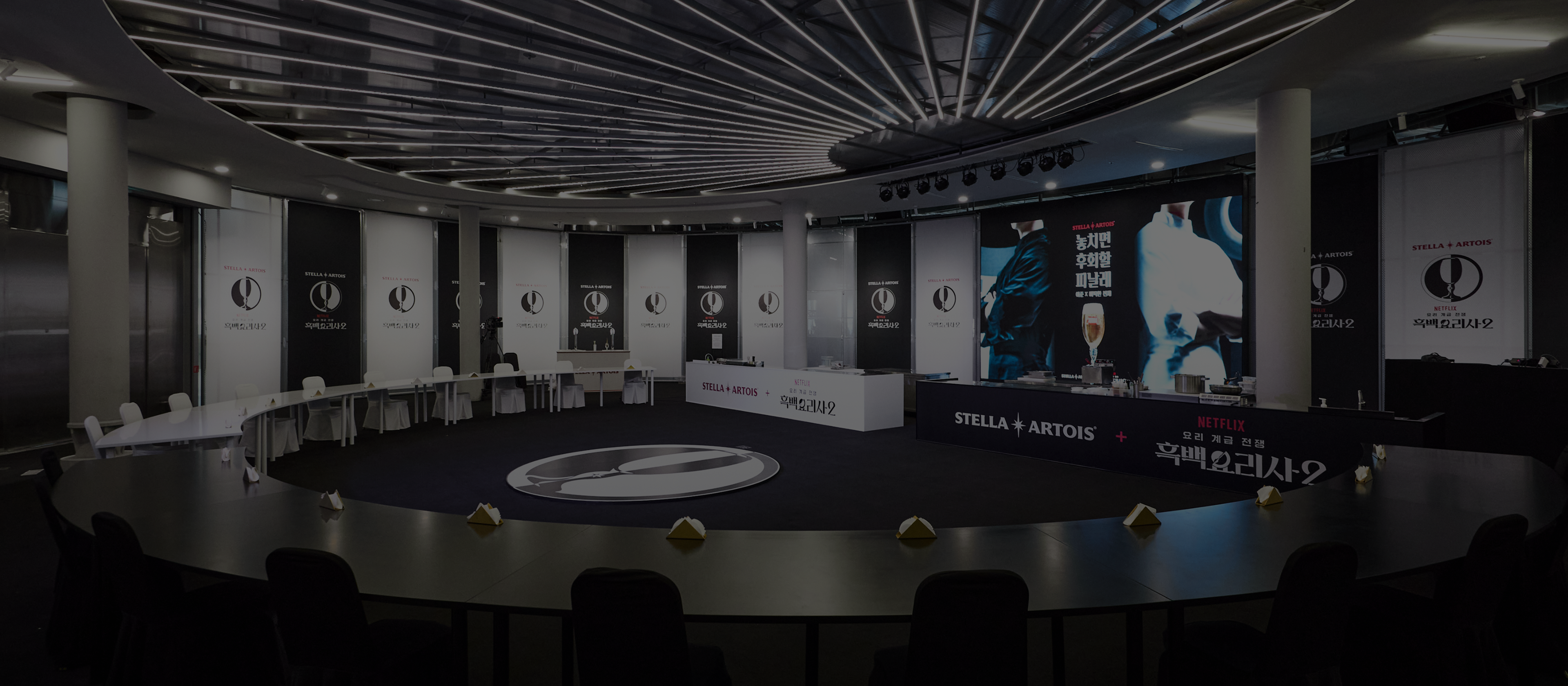 어두운 조명 아래 원형 테이블이 배치된 넓은 이벤트 홀입니다. 스텔라 아르투아와 흑백요리사 로고가 새겨진 대형 배너들이 무대 주변을 둘러싸고 있으며 중앙에는 스텔라 아르투아 로고가 빛나고 있습니다